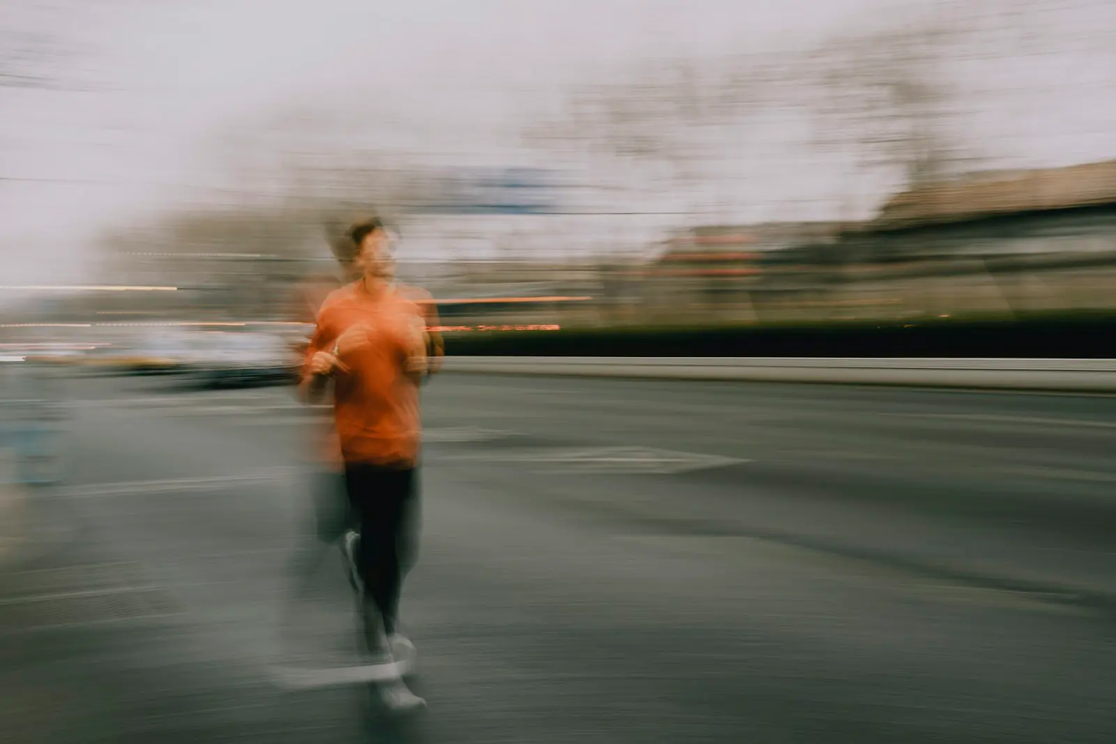 A motion-blurred person in an orange shirt and dark trousers running on a wide road, with bare trees and traffic behind.