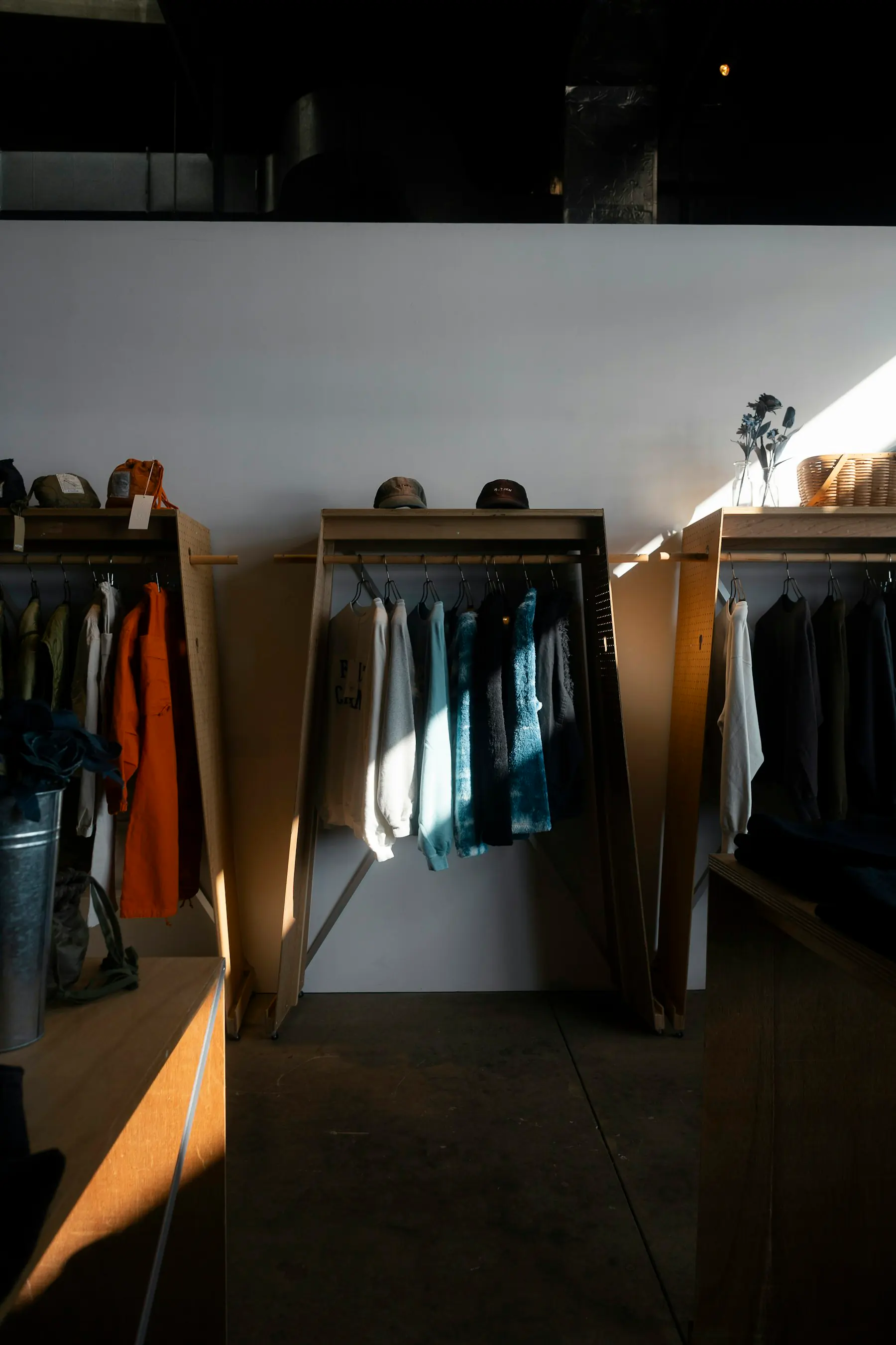 Three wooden clothing racks in a sunlit shop, holding jackets, sweatshirts, and shirts, with caps on the center rack.