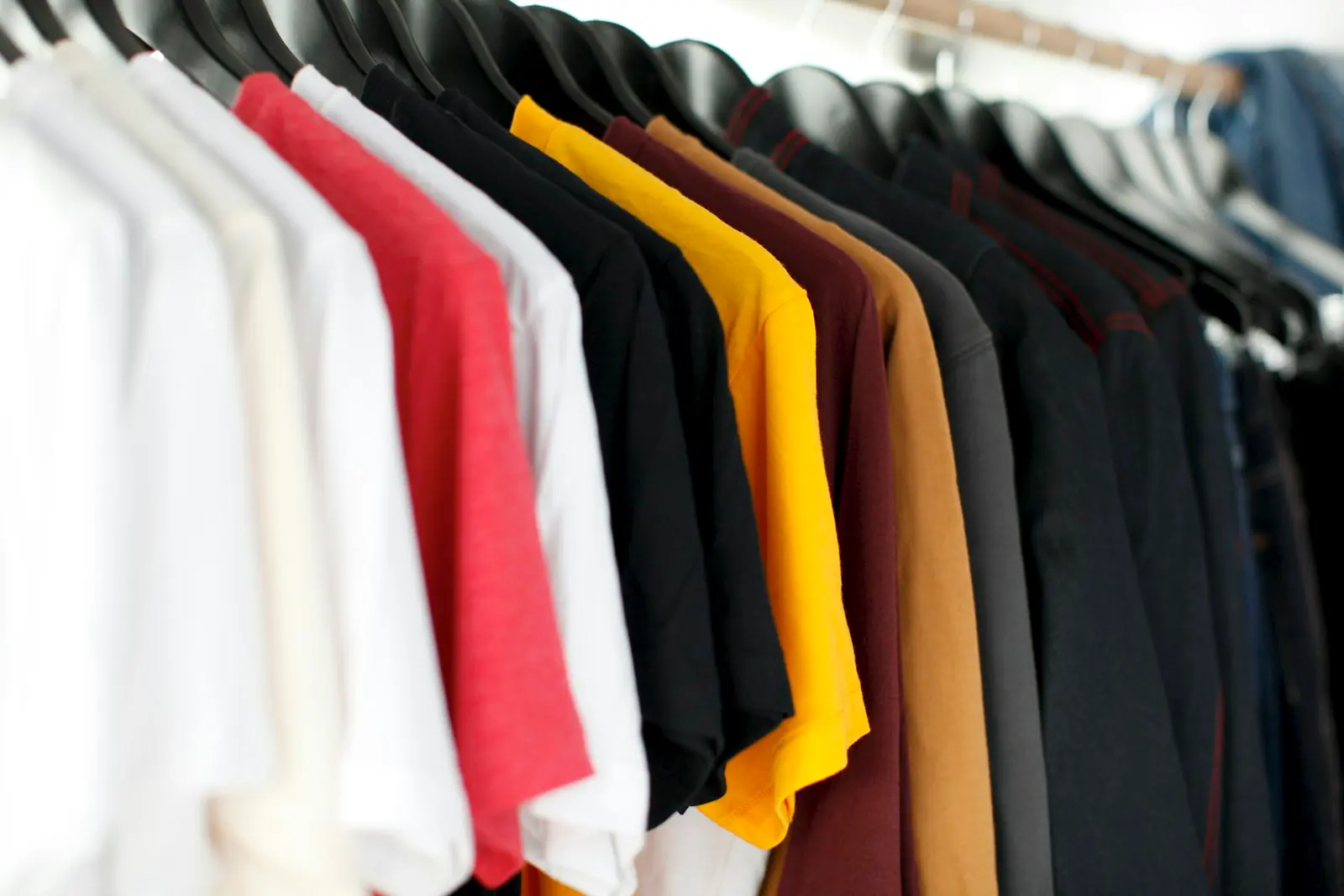 A row of short-sleeve t-shirts in white, red, black, yellow, brown, and navy hanging on black hangers.