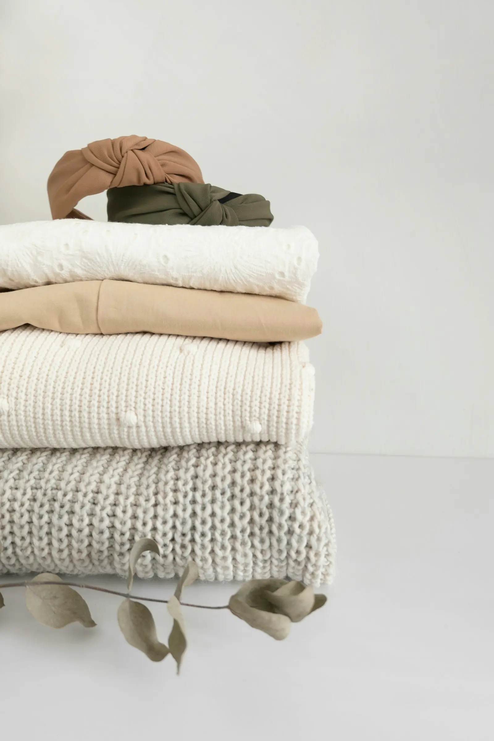 A stack of folded cream, beige, and grey knitwear with two knotted fabric headbands on top, on a white surface.