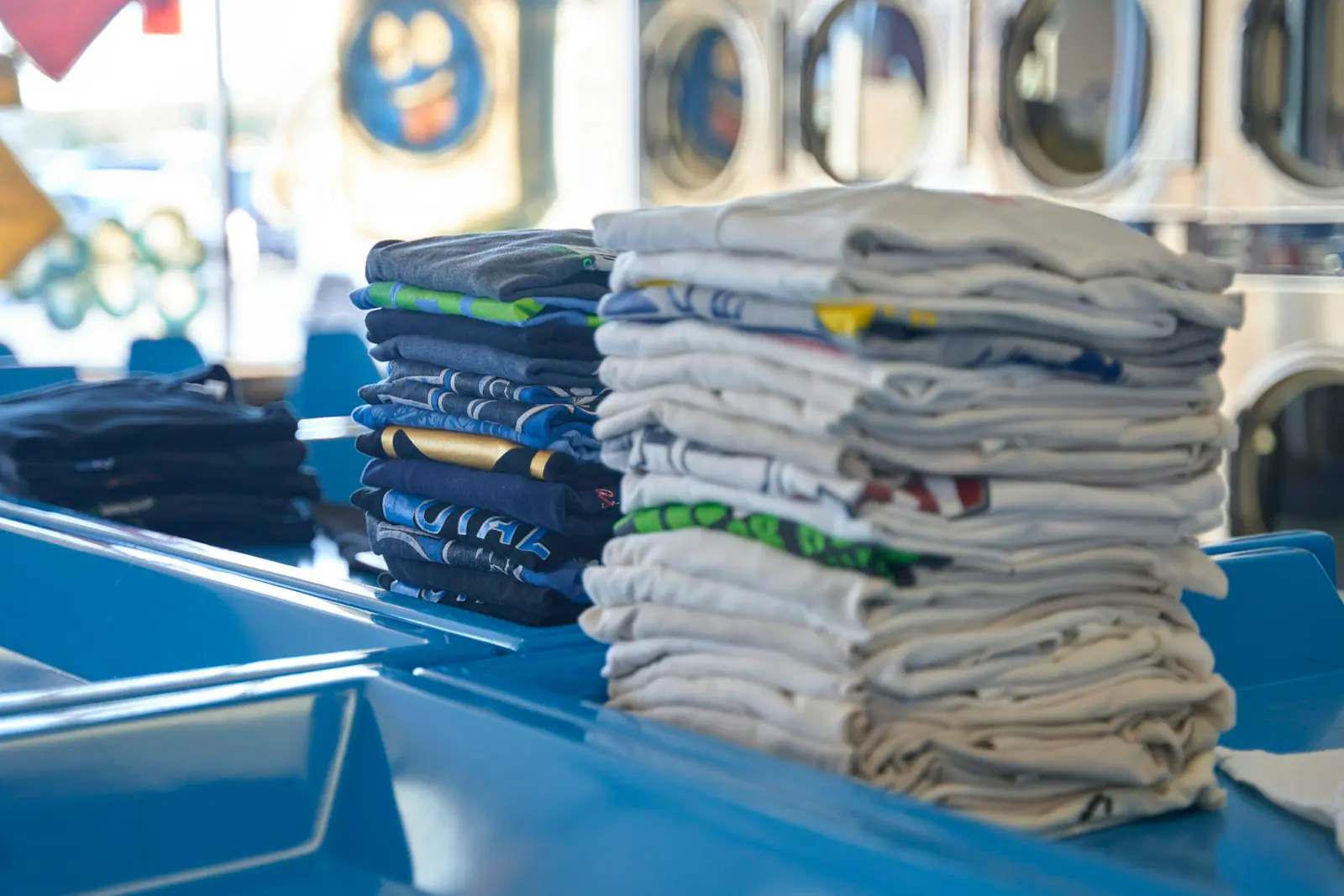 Stacks of folded graphic and plain t-shirts on a blue counter, with washing machines visible behind them.